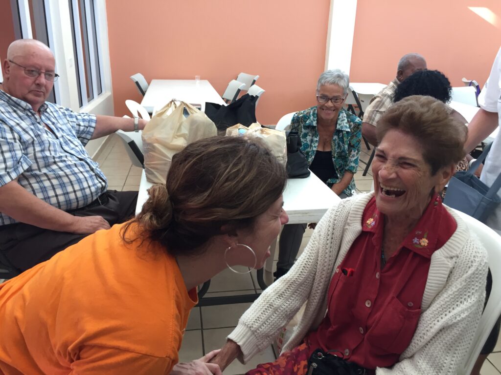 Danielle Butin and an older woman in a red shirt and white cardigan are laughing together. Older adults are smiling in the background.